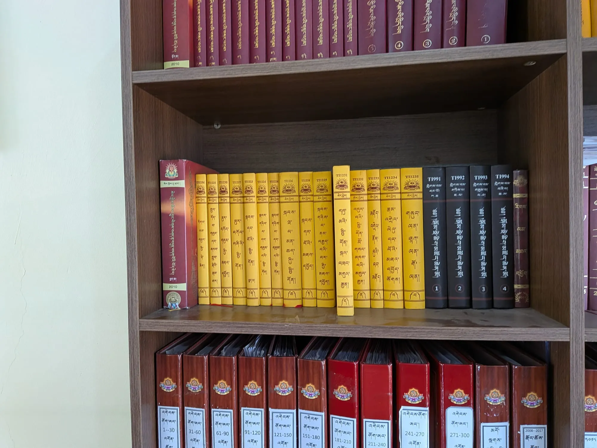 A library shelf holding fifteen yellow volumes, each bearing the Sera Jey emblem, catalogued TT1221 to TT1236, alongside darker bound volumes and archive files.
